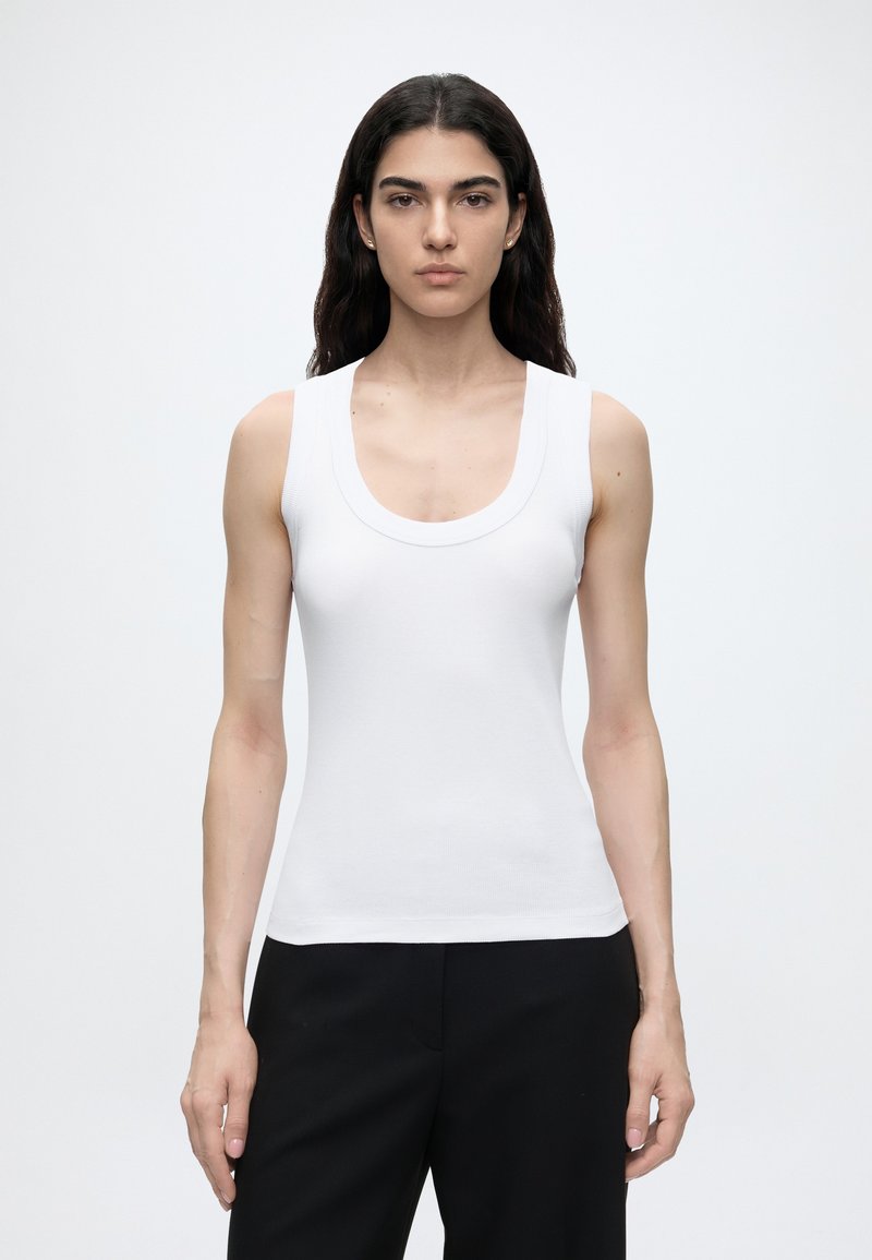 Femme aux longs cheveux foncés portant un haut blanc sans manches et un pantalon noir, debout devant un fond blanc uni.