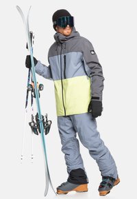 Skis påklædning med en grå og limegrøn jakke, lysegrå bukser, sorte handsker og skibriller. Holder på blå og hvide ski med sorte bindinger.