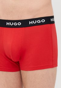 Piros pamut boxer alsó, fekete derékpánttal, amelyen a "HUGO" fehér logó található. Az anyag sima textúrájú és minimális varrással rendelkezik.