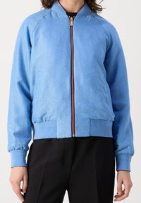 Chaqueta bomber azul con textura paisley, puños y dobladillo de canalé, y una cremallera frontal de herraje dorado. Lleva puesta sobre pantalones negros.