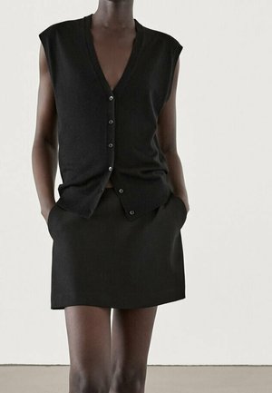 Ærmeløs sort vest med knapper kombineret med en sort mininederdel, model stående med hænderne i nederdelslommerne mod en ensfarvet baggrund.