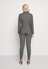 Costume gris sur mesure avec un blazer cintré et un pantalon slim. Le tissu présente un motif à carreaux subtil. Accessoirisé avec des sandales à talons noires.