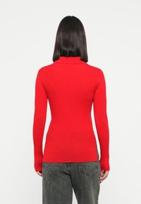Pull à col roulé rouge côtelé avec des manches longues, design ajusté et texture lisse, associé à un jean foncé ample.