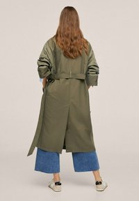 Olivově zelený trench coat s páskem v pase, širokými rukávy a ventilačním otvorem na zadní straně, v kombinaci s širokými denimovými culotty a světlými botami.