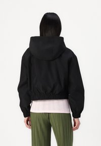 Black cropped jacket with a hood, elastic waistband, and long sleeves. Worn over a light pink textured top and green pleated pants.