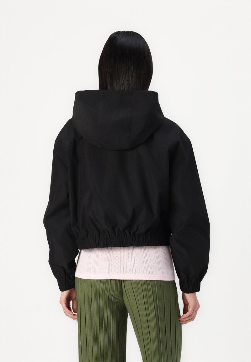 Giacca nera corta con cappuccio, vita elastica e maniche lunghe. Indossata sopra un top texturizzato rosa chiaro e pantaloni verdi plissettati.