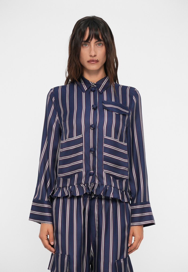 Blusa a righe blu navy e crema con maniche lunghe, chiusura a bottoni, orlo arricciato e taschino sul petto. Tessuto liscio e leggero.