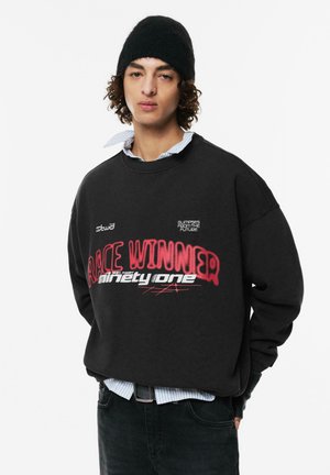 Schwarzer, übergroßer Sweatshirt mit grafischen Drucken, die "RACE WINNER" und "einundneunzig" lesen. Darüber getragen ist ein hellblaues Hemd mit Kragen.