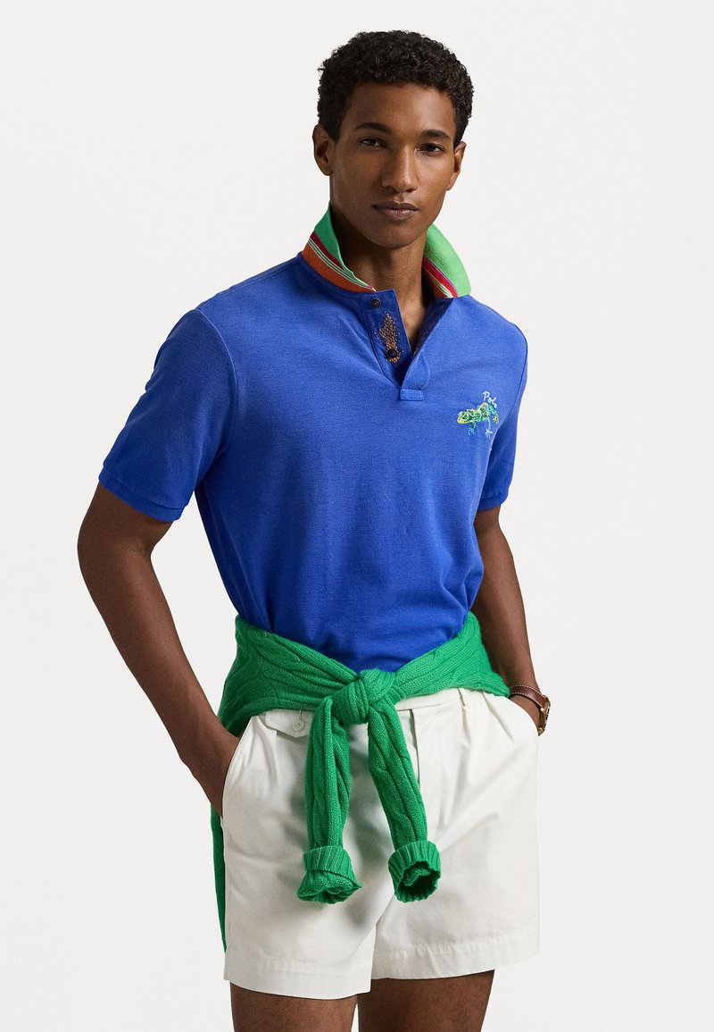 Camisa polo azul com colarinho listrado em verde e vermelho, detalhe bordado no peito, combinada com calções brancos e um suéter verde amarrado à cintura.