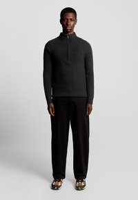 Donkergrijze geribbelde gebreide trui met een hoge kraag en een halve rits, gecombineerd met losse zwarte broeken en gestructureerde schoenen. Over het algemeen een soepele textuur.