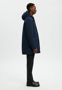 Cappotto imbottito blu navy con cappuccio, maniche lunghe e taglio dritto, abbinato a jeans scuri e scarpe nere, visto di lato.