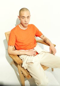 T-shirt orange à manches courtes avec col blanc, associé à un pantalon beige clair. Sujet assis sur une chaise en bois tissée avec une posture détendue.