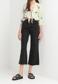 Kvinde iført en blomstret bluse med knyt foran, højtaljede sorte cropped brede jeans og sorte sandaler med nitter.