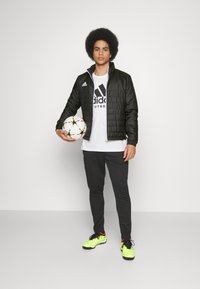 Sort quiltet jakke, hvid Adidas t-shirt, sorte bukser og lysegrønne fodboldstøvler. Holder en hvid fodbold med multikolor accent.