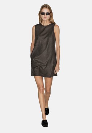 Donna che cammina indossando un abito mini senza maniche grigio scuro, scarpe slip-on nere e occhiali da sole rettangolari neri su uno sfondo bianco.