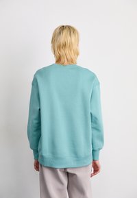 Persona con cabello rubio corto, vistiendo una sudadera teal holgada y pantalones beige claros, de pie frente a una pared blanca lisa.