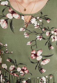 Vestido floral verde con escote redondeado y detalle en forma de ojal, flores negras y rosas, y una cintura fruncida. Incluye collares dorados en capas.