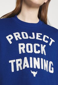 Blå sweatshirt med "PROJECT ROCK TRAINING" i stora vita slitna bokstäver. Rund halsringning och mjuk tygtextur.