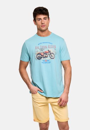 Hombre con camiseta azul claro con gráfico de motocicleta vintage y pantalones cortos amarillos remangados, de pie con una mano en el bolsillo.