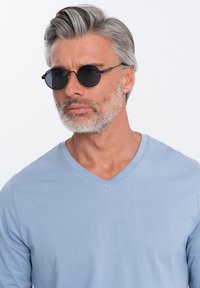 Hellblaues langärmliges V-Ausschnitt-Shirt aus weichem Stoff, kombiniert mit runden schwarzen Sonnenbrillen, die das Gesicht und die Merkmale klar betonen.