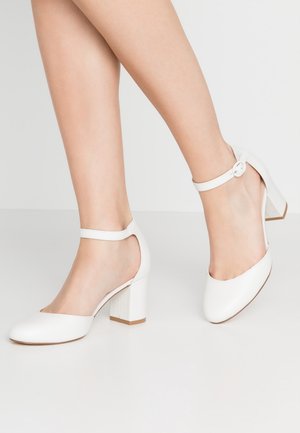 Beine mit weißen High Heels mit Knöchelriemen und abgerundeten Zehen auf einer hellen einfarbigen Oberfläche und Hintergrund.