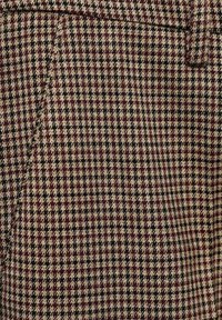 Tissu en mélange de laine avec un motif pied-de-poule texturé en bordeaux, beige et noir. Présente une coupe ajustée avec des coutures précises.