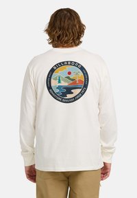 Mann mit welligem Haar, der ein weißes Langarmhemd mit einem runden Billabong-Logo trägt, das auf der Rückseite einen Berg, eine Palme und einen Fluss zeigt.