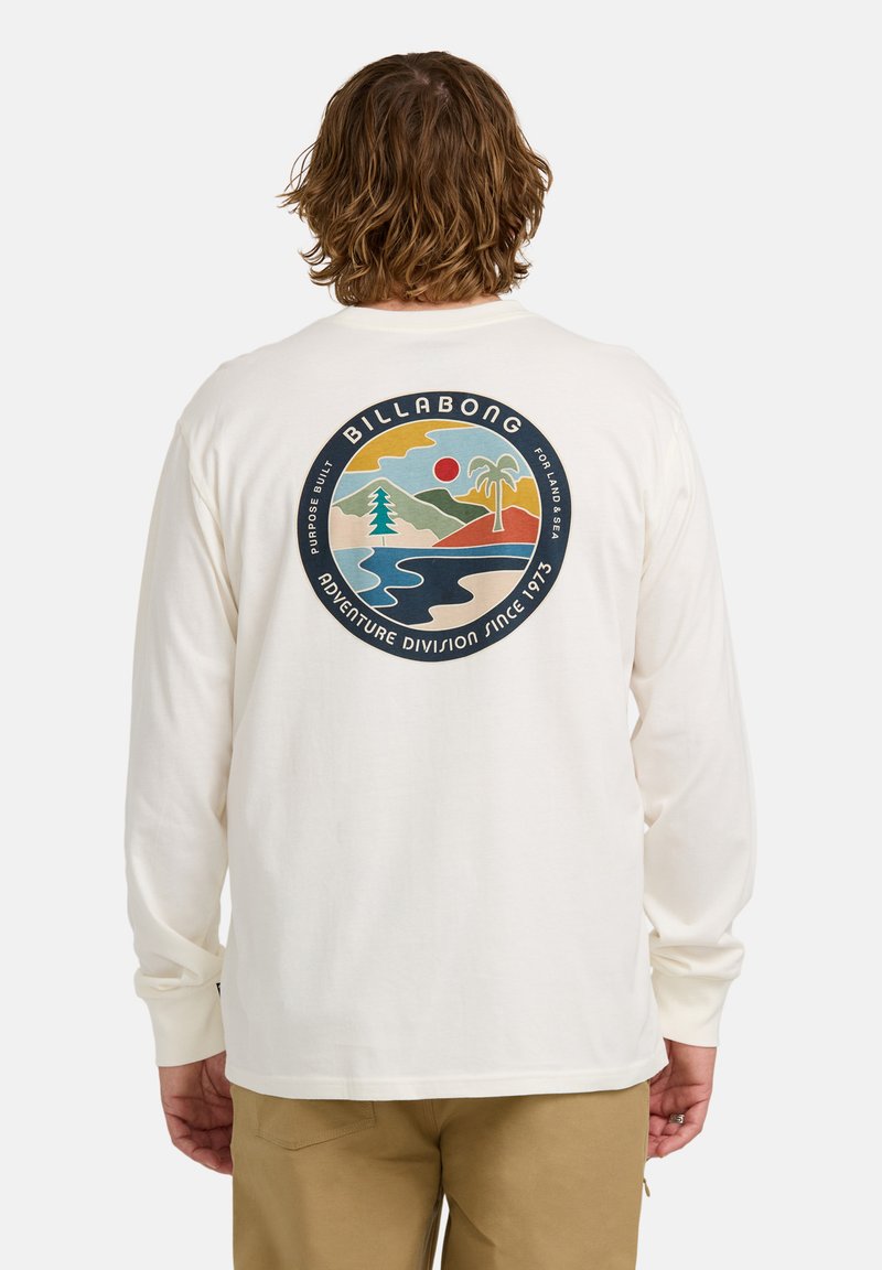 Mann mit welligem Haar, der ein weißes Langarmhemd mit einem runden Billabong-Logo trägt, das auf der Rückseite einen Berg, eine Palme und einen Fluss zeigt.