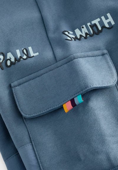 Zila sporta jumprene ar nosaukumiem "PAUL" un "SMITH" kontrastējošos materiālos. Iekļauta kabata ar atloku un krāsains svītrains akcents.