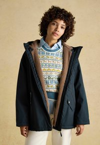 Námornícky modrý zimný kabát s umelým kožušinovým podšívkou, veľkou kapucňou, vreckami na zips, cez viacfarebný vzorovaný sveter a biele nohavice.