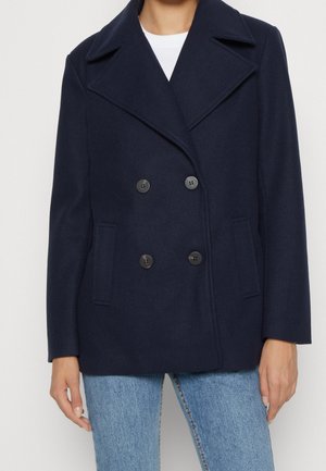 Kobieta w granatowym dwurzędowym płaszczu typu pea coat z szerokimi klapami i przednimi kieszeniami, w połączeniu z jasnoniebieskimi jeansami.