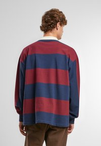 Košeľa s dlhými rukávmi s hlbokými červenými a námorníckymi horizontálnymi pruhmi. Vyrobená z mäkkého materiálu s voľným strihom a výstrihom do okrúhla.