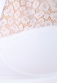 Sostén blanco con detalles de encaje en el borde superior, que presenta una textura de tela suave y una forma curva, destacando intrincados patrones de encaje floral.