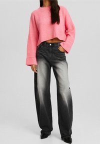 Femme portant un pull court rose et un jean noir taille haute à jambes larges avec des détails gris délavés, debout avec une main dans la poche.