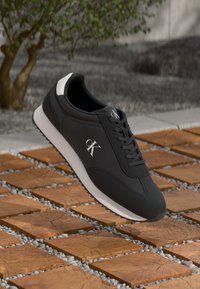 Black athletic shoe with a sleek design, featuring a white heel accent and a textured rubber sole. The logo is displayed on the side.