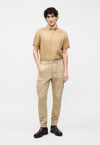 Άντρας με σγουρά σκούρα μαλλιά στέκεται, φορώντας καφέ πουκάμισο με κοντά μανίκια και κουμπιά, μπεζ παντελόνι cargo και σκούρα καφέ μπότες, σε λευκό φόντο.