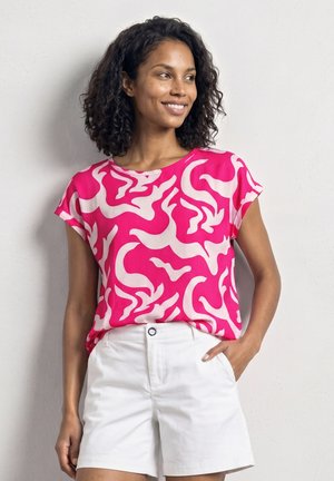 Vrouw met krullend haar die lacht, draagt een roze en witte abstracte korte mouw top en witte korte broek, staand tegen een effen muur.