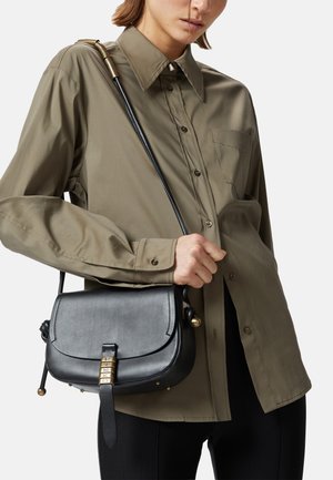 Bolso de hombro de cuero negro con forma curvada, herrajes dorados y cierre de broche; combinado con una camisa de botones verde oliva.