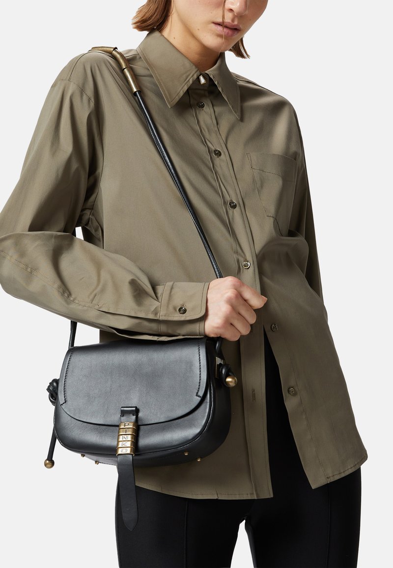Borsa a tracolla in pelle nera con forma curva, accessori in metallo dorato e chiusura a scatto; indossata con una camicia verde oliva a bottoni.