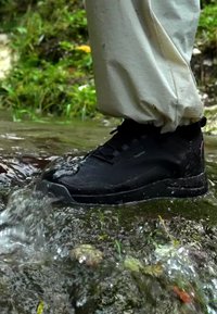 Černá voděodolná bota vstupuje do mělké tekoucí vody na lesní cestě s světlými kalhotami.