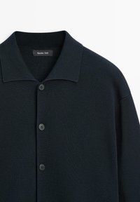 Navy blue knit shirt with a short collar, featuring four black buttons down the front, textured fabric, minimalistic design.