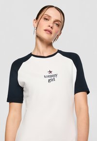 Hvit t-skjorte med marineblå raglanermer, med "tommy girl" brodert i marineblått og rødt på brystet. Glatt tekstur, uformelt design.
