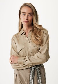 Camicia lunga manica color beige chiaro con chiusura a bottoni e due tasche sul petto. Tessuto morbido con una vestibilità rilassata, abbinata a pantaloni a vita alta.