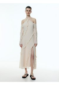 Lang, beige off-shoulder jurk met gerimpelde accenten, een zijsplit en doorschijnende stof. Gekleed met zwarte platform slippers.