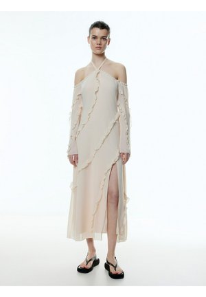 Lang, beige off-shoulder jurk met gerimpelde accenten, een zijsplit en doorschijnende stof. Gekleed met zwarte platform slippers.