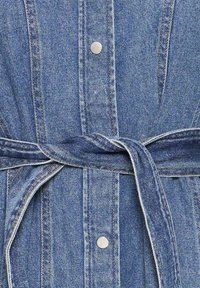 Vêtement en denim bleu avec des coutures verticales, des boutons-pression argentés et une ceinture nouée serrant la taille.