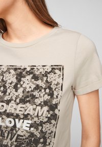 Béžové tričko s krátkým rukávem a kulatým výstřihem. Obsahuje černobílý květinový grafický potisk a text "SNĚJ. MILUJ. ŽIJ."