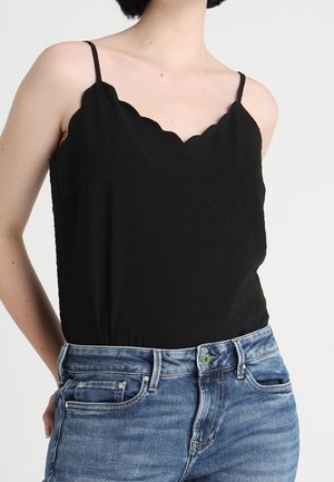 Kobieta w czarnym, bezrękawnikowym topie z falistym brzegiem i cienkimi ramiączkami, włożonym w wyblakłe niebieskie dżinsy.