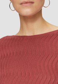 Rotes gestricktes Oberteil mit wellenförmiger Textur und weitem Ausschnitt. Sichtbarer silberner Creolenohrring, mit einem glatten Hauthintergrund.