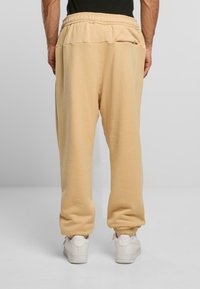 Bētējo sporta bikses ar elastīgu jostu, sa�šaurinātiem piedurknēm un brīvu piegriezumu. Valkātas ar baltām trenažierēm, parādītas no aizmugures.